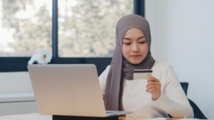 Seorang wanita sedang memegang kartu kredit dan bekerja di laptop, relevan dengan konsep Deposito Syariah Menurut Islam dalam keuangan digital.