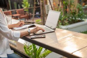 Seorang wanita sedang mengetik di laptop sambil menulis di buku catatan di meja kafe luar ruangan, dengan ponsel di sampingnya.