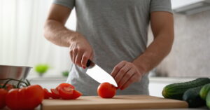 Seorang pria sedang memotong tomat dengan pisau di dapur, dengan bahan makanan lain di atas meja.