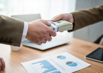 Dua pria bertukar uang tunai dalam jumlah besar di atas meja kantor dengan latar belakang laptop dan dokumen grafik keuangan.