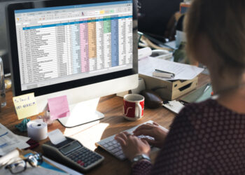 Seseorang sedang bekerja dengan Template Keuangan Excel di layar komputer sambil menikmati secangkir kopi di meja kantor.