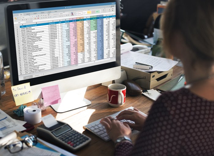 Seseorang sedang bekerja dengan Template Keuangan Excel di layar komputer sambil menikmati secangkir kopi di meja kantor.