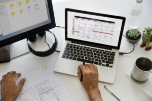 Seseorang sedang merencanakan jadwal di laptop dengan kalender digital dan catatan kerja di meja kerja.