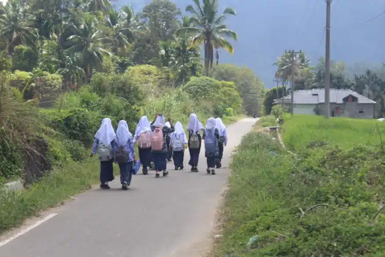 Rombongan siswi berseragam sekolah dan berjilbab putih berjalan pulang di jalan desa dekat sawah, tampak dari belakang