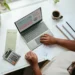 Tampak atas orang memeriksa tabel keuangan di laptop dengan kalkulator, lembar dokumen, dan amplop di meja