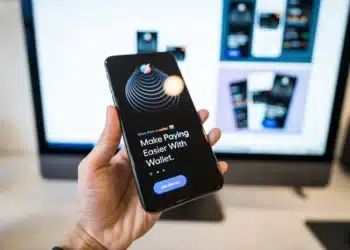 Tangan memegang ponsel yang menampilkan aplikasi dompet digital dengan layar monitor buram di latar belakang