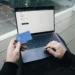 Orang duduk dengan laptop sambil memegang kartu kredit