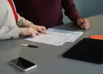 Dua orang memeriksa lembaran formulir di atas meja, ponsel dan tablet di dekatnya