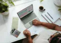 Tampak atas orang memeriksa tabel keuangan di laptop dengan kalkulator, lembar dokumen, dan amplop di meja