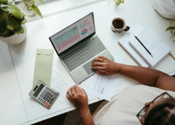 Tampak atas orang memeriksa tabel keuangan di laptop dengan kalkulator, lembar dokumen, dan amplop di meja