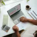 Tampak atas orang memeriksa tabel keuangan di laptop dengan kalkulator, lembar dokumen, dan amplop di meja