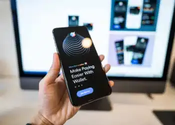 Tangan memegang ponsel yang menampilkan aplikasi dompet digital dengan layar monitor buram di latar belakang
