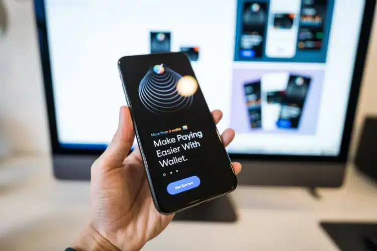 Tangan memegang ponsel yang menampilkan aplikasi dompet digital dengan layar monitor buram di latar belakang