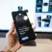 Tangan memegang ponsel yang menampilkan aplikasi dompet digital dengan layar monitor buram di latar belakang