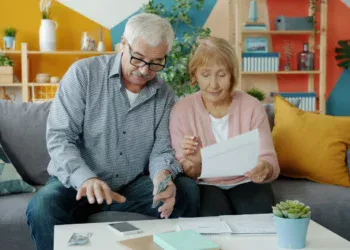Pasangan suami istri lanjut usia memeriksa dokumen keuangan dan uang tunai di sofa, gambaran pemilik rekening gabungan