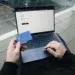 Orang duduk dengan laptop sambil memegang kartu kredit